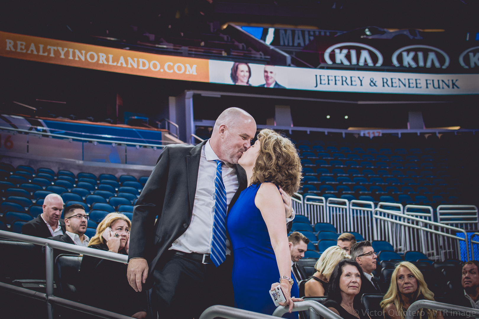 Jeff and Renee Funk share a kiss in the stands at Amway Center as their sponsor signage glows behind them.